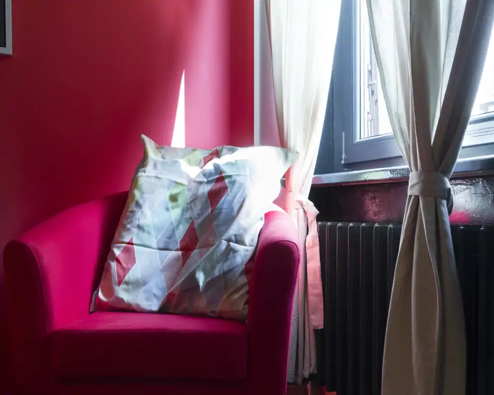 Un angolo accogliente con una poltrona rosa brillante e un cuscino decorativo, posizionata accanto a una finestra luminosa con tende neutre.