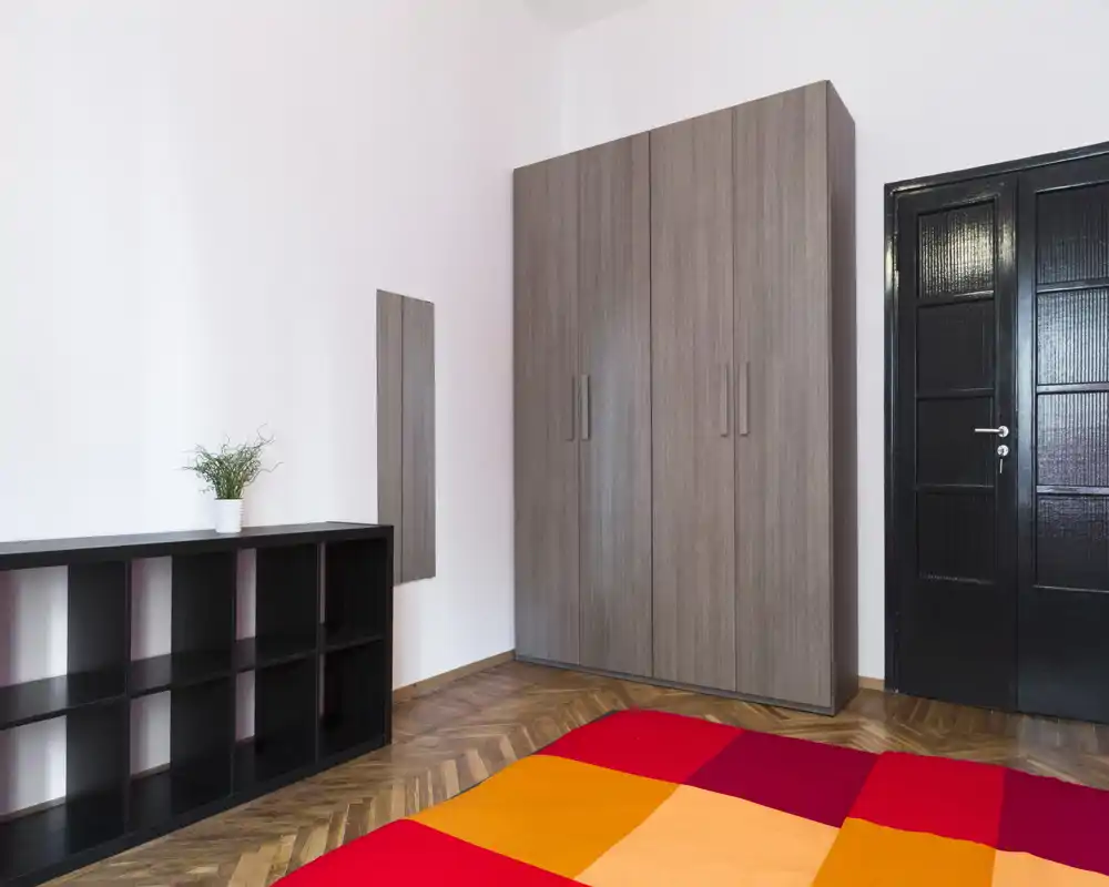 Camera da letto luminosa e minimal con grande armadio, scaffalatura bassa e una coperta colorata; pavimento in parquet e arredo essenziale per un aspetto moderno e pulito.