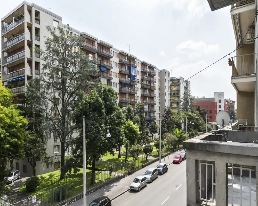 Vista dal balcone su una strada alberata e condomini, con auto parcheggiate e un piccolo spazio verde.
