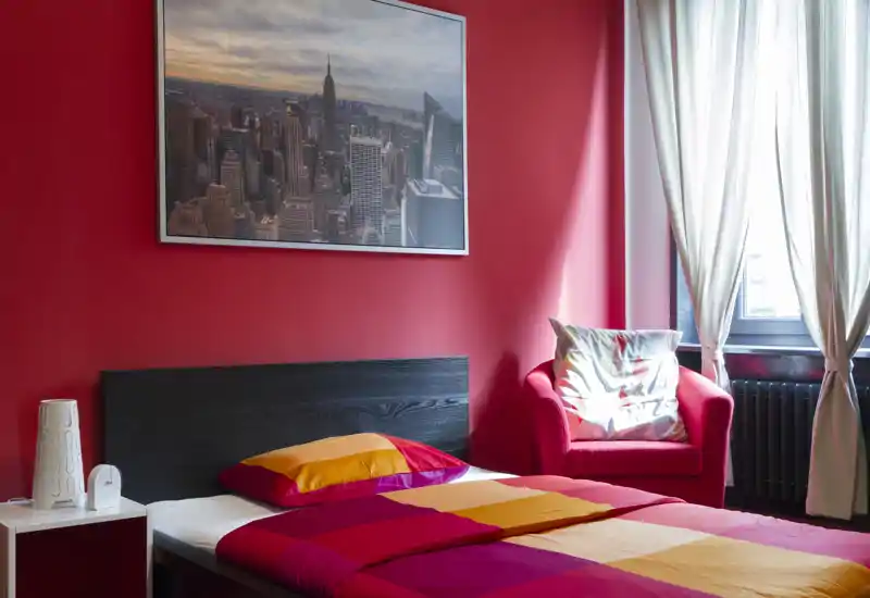 Camera da letto luminosa e colorata con letto matrimoniale contro una parete rossa e una poltrona comoda vicino alla finestra. Biancheria vivace, quadro con città e luce naturale soffusa.