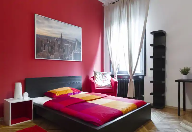 Camera da letto moderna e luminosa con letto basso su piattaforma scura, copriletto colorato e parete rossa d'accento; ampia finestra e una poltrona confortevole rendono l'ambiente accogliente.