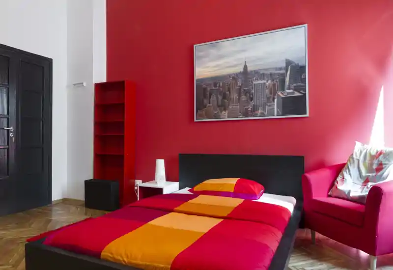 Camera da letto luminosa e vivace con letto matrimoniale, testiera moderna e una poltrona accogliente vicino alla finestra. Parete rossa e quadro con scorcio cittadino donano personalità.