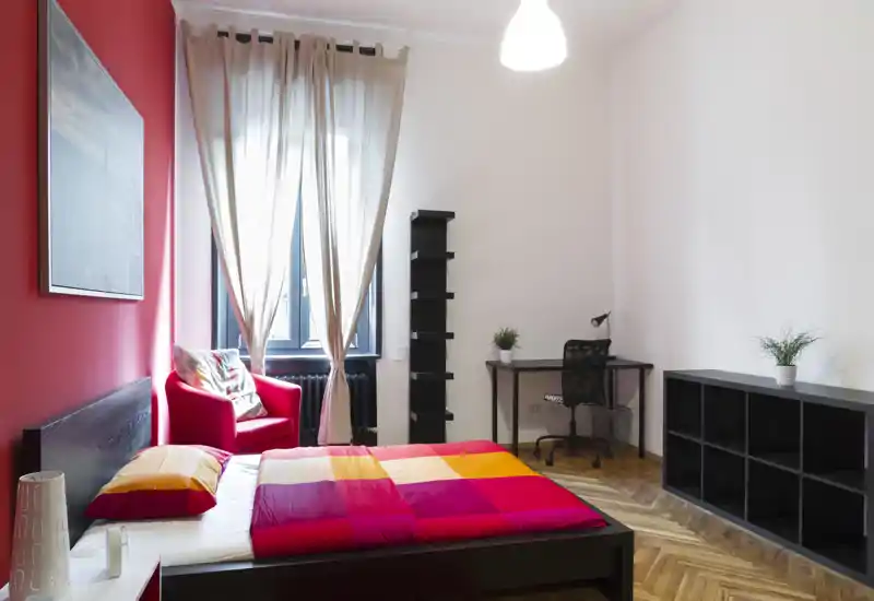 Camera da letto moderna e luminosa con un letto matrimoniale colorato, parete rossa e una poltrona accogliente vicino alla finestra. Angolo studio e scaffalature rendono la stanza adatta sia al riposo che al lavoro leggero.