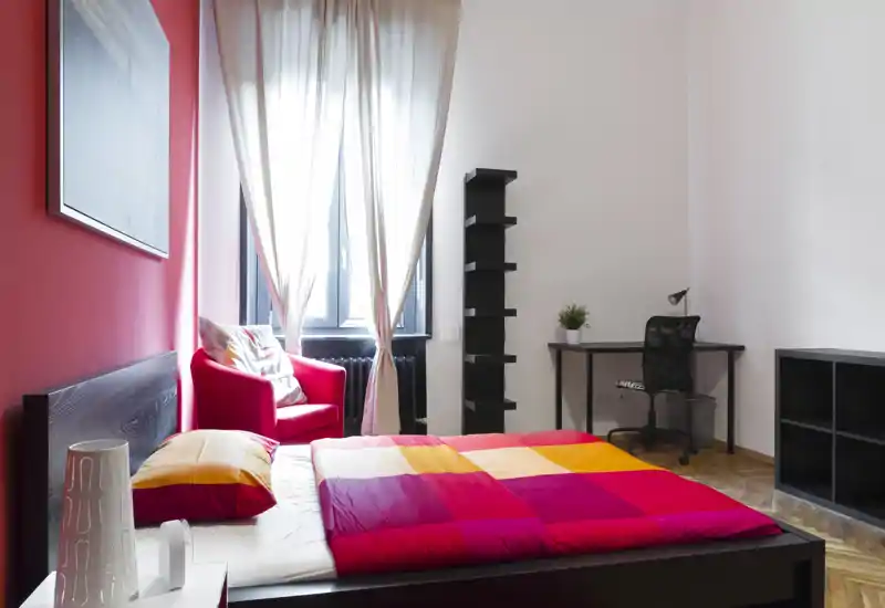 Camera da letto moderna e luminosa con letto matrimoniale colorato, parete rossa e una poltrona vicino alla finestra; scrivania e scaffali piccoli rendono lo spazio funzionale.