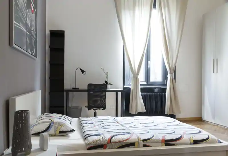 Camera da letto moderna e luminosa con biancheria a fantasia, scrivania vicino alla finestra e luce naturale soffusa — ideale per un soggiorno confortevole.