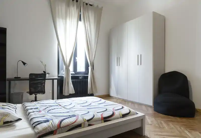 Camera da letto luminosa e moderna con letto matrimoniale con copriletto a fantasia, ampia finestra con tende chiare, armadio e una piccola scrivania — ideale per soggiorni confortevoli.