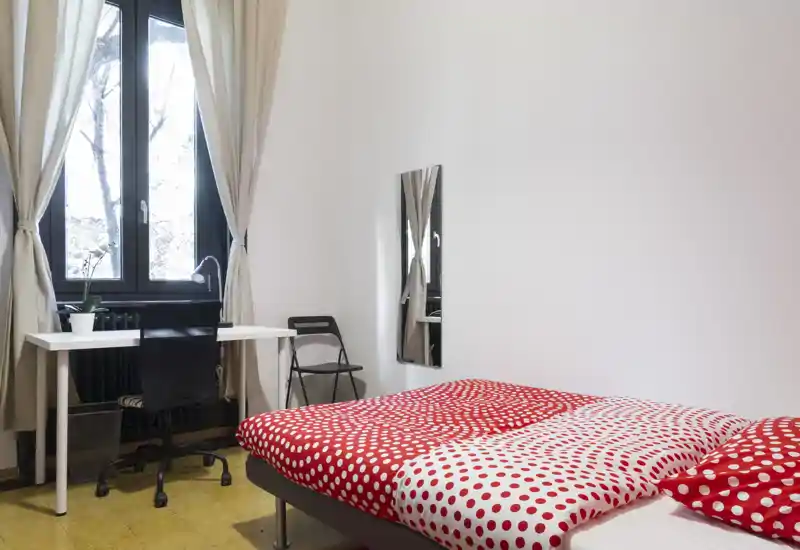 Camera da letto luminosa e ordinata con letto matrimoniale coperto da una trapunta a pois rossi e bianchi, una scrivania vicino alla finestra e arredi essenziali e moderni.