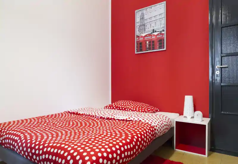 Camera singola luminosa e di stile con copripiumino rosso a pois, mensolina come comodino e parete d'accento rossa — accogliente e moderna per un soggiorno in solitaria.