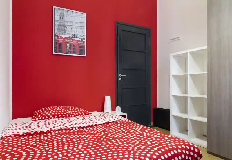 Camera da letto compatta e moderna con parete rossa a contrasto e biancheria a pois; presente comodino e mobile a cubi, luminosa e di design.