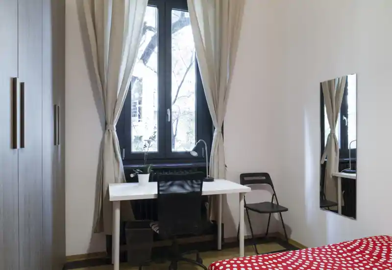 Piccola camera da letto con angolo di letto dal copriletto rosso a pois, una scrivania bianca davanti a una finestra alta con tende beige, una sedia da ufficio nera e un armadio — luminosa e arredata in modo essenziale, ideale per una persona.