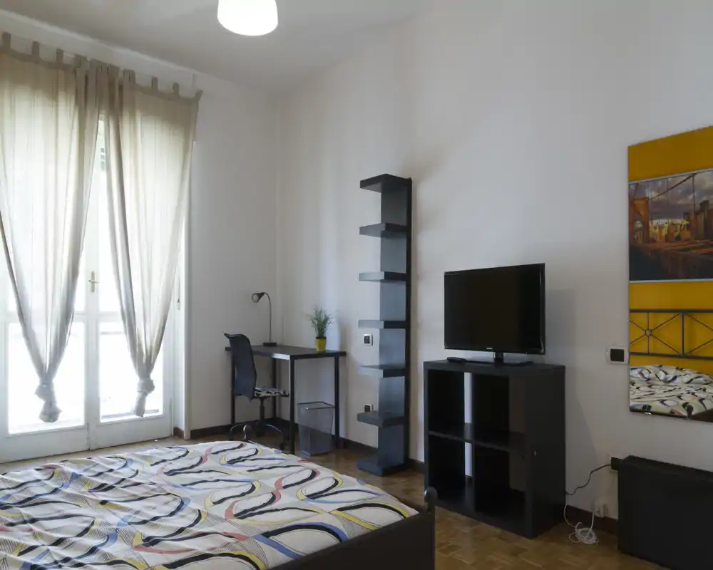 Camera da letto moderna e luminosa con ampio letto, biancheria a fantasia, scrivania vicino alla finestra e mobile porta TV — ideale per single o coppie che cercano una stanza elegante e ben illuminata.