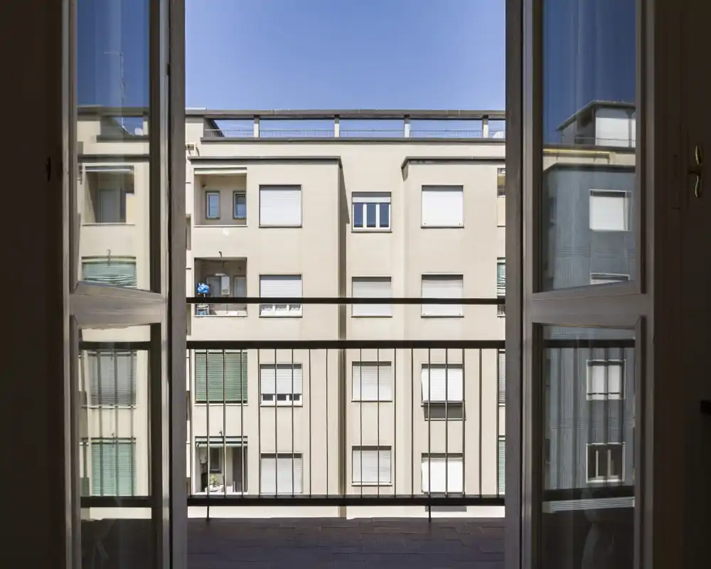 Piccolo balcone visto attraverso porte doppie aperte, con affaccio sulle facciate degli appartamenti vicini sotto luce diurna chiara.