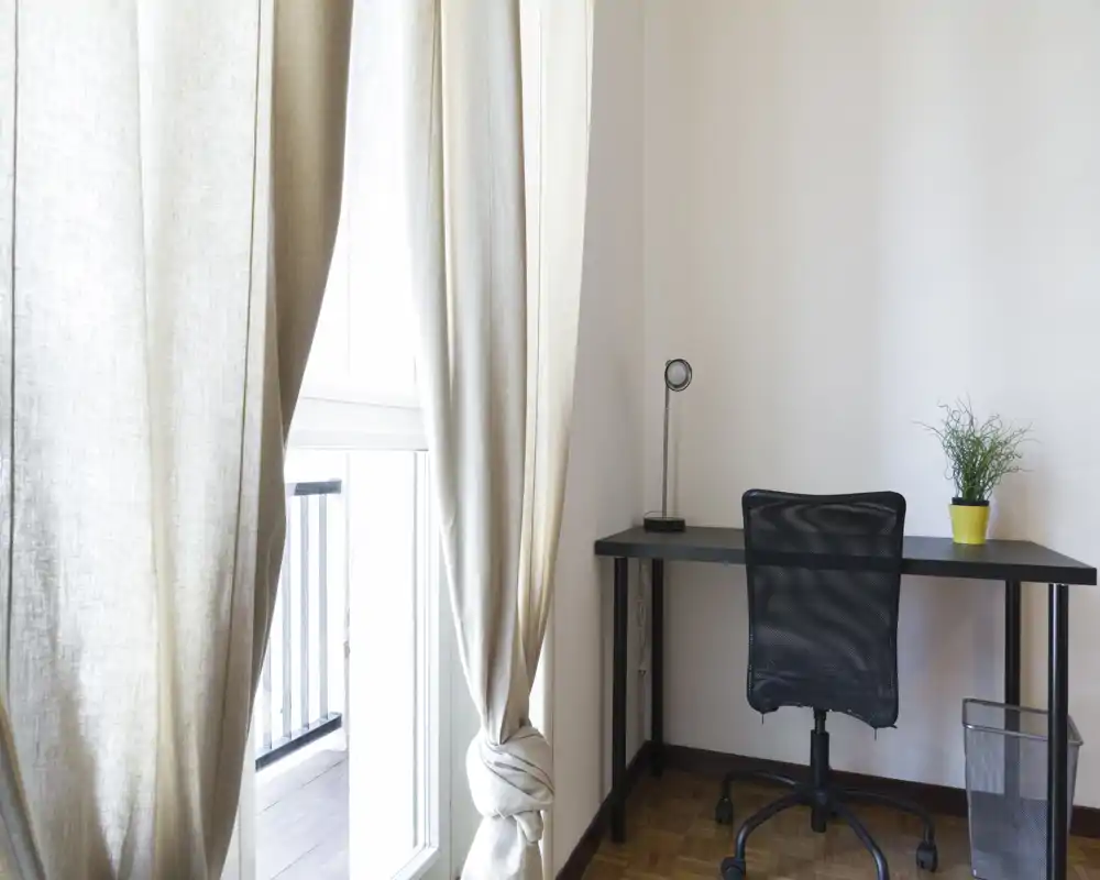 Angolo studio luminoso e minimal con scrivania nera, sedia in mesh e pianta in vaso accanto a una grande finestra con tende in lino chiare.