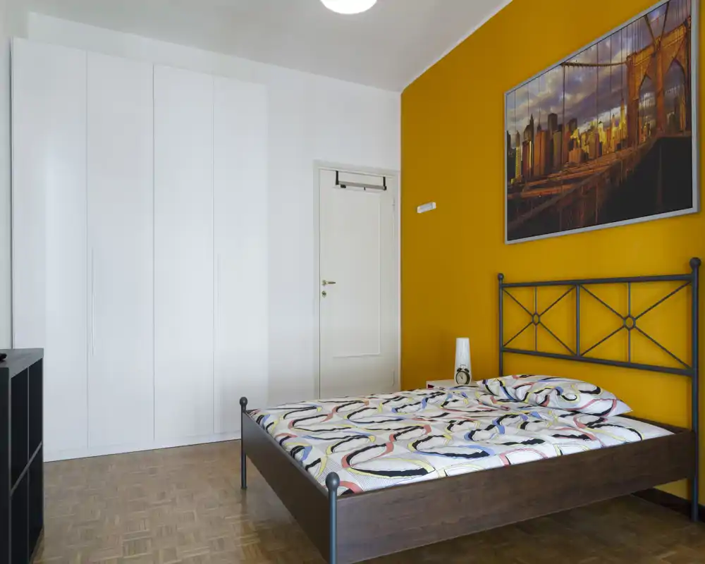 Camera da letto moderna e luminosa con letto matrimoniale con struttura in metallo, biancheria a fantasia e un grande armadio. Una parete d'accento gialla e un quadro incorniciato donano un tocco contemporaneo.