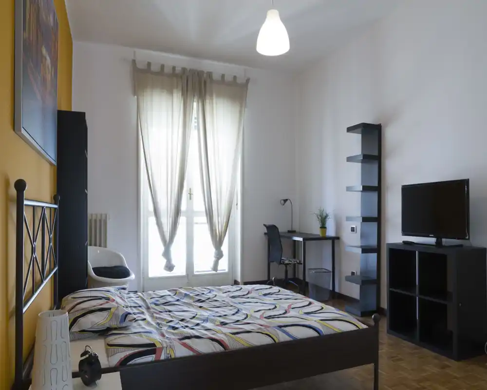 Camera da letto luminosa e ben arredata con letto matrimoniale, scrivania e mobile TV; la luce naturale filtrata dalle tende crea un'atmosfera accogliente.