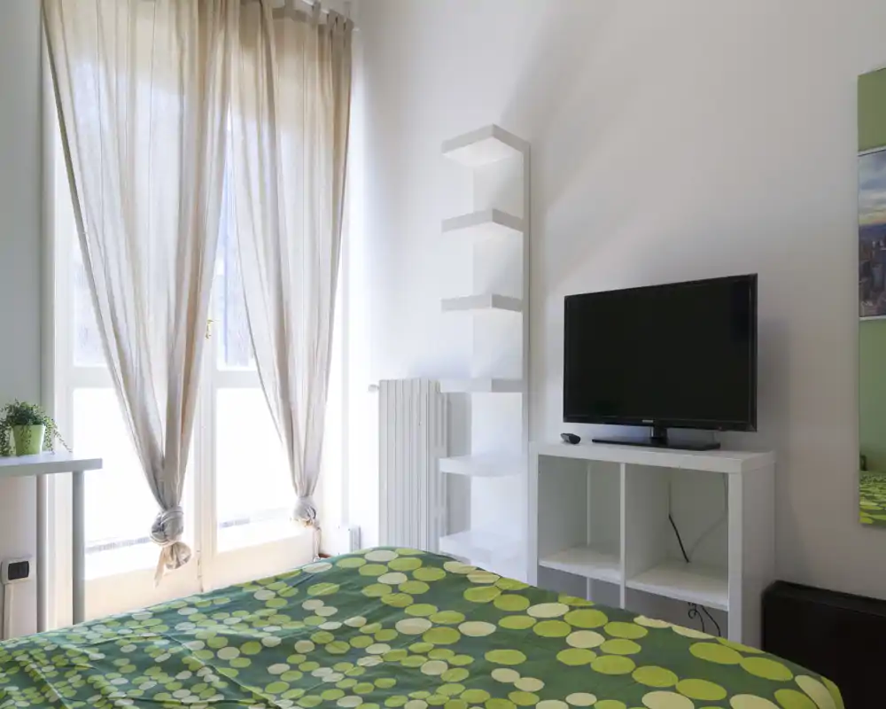 Camera da letto luminosa e minimal con copriletto verde a fantasia, ampia finestra con tende chiare, TV su mobile bianco e una mensola alta accanto al termosifone.