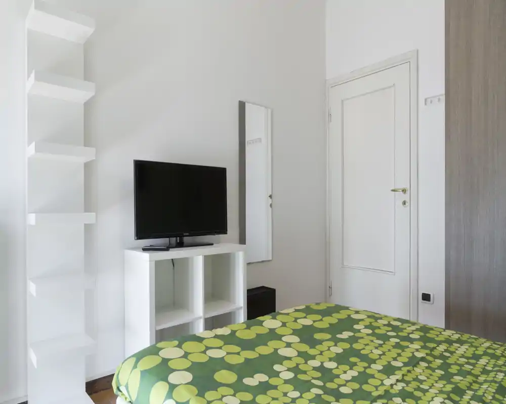 Camera da letto luminosa e moderna con copriletto verde a fantasia, specchio a parete e TV su un mobiletto bianco basso. Lo spazio è ordinato e semplicemente arredato con buona luce naturale.