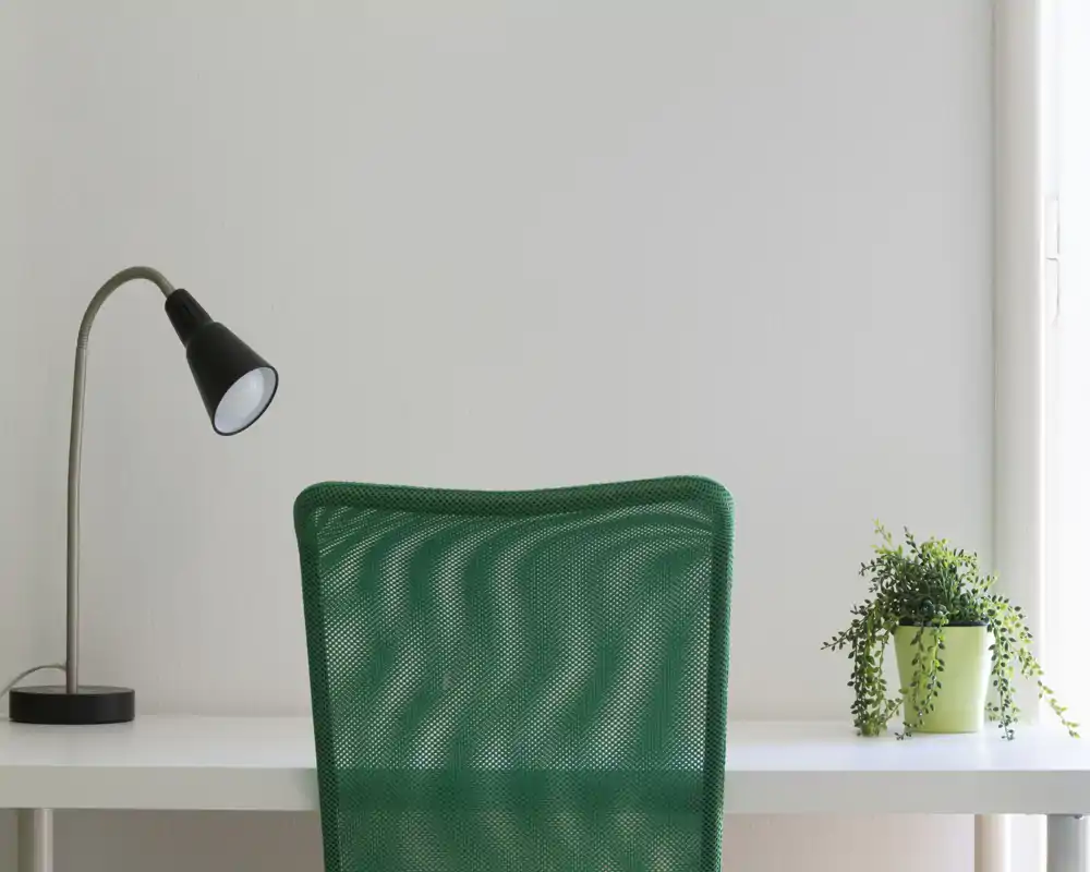 Angolo scrivania minimal e luminoso con una sedia da ufficio verde in rete, una lampada da tavolo nera e una piccola pianta in vaso — un'area ordinata ideale per lavoro o studio.
