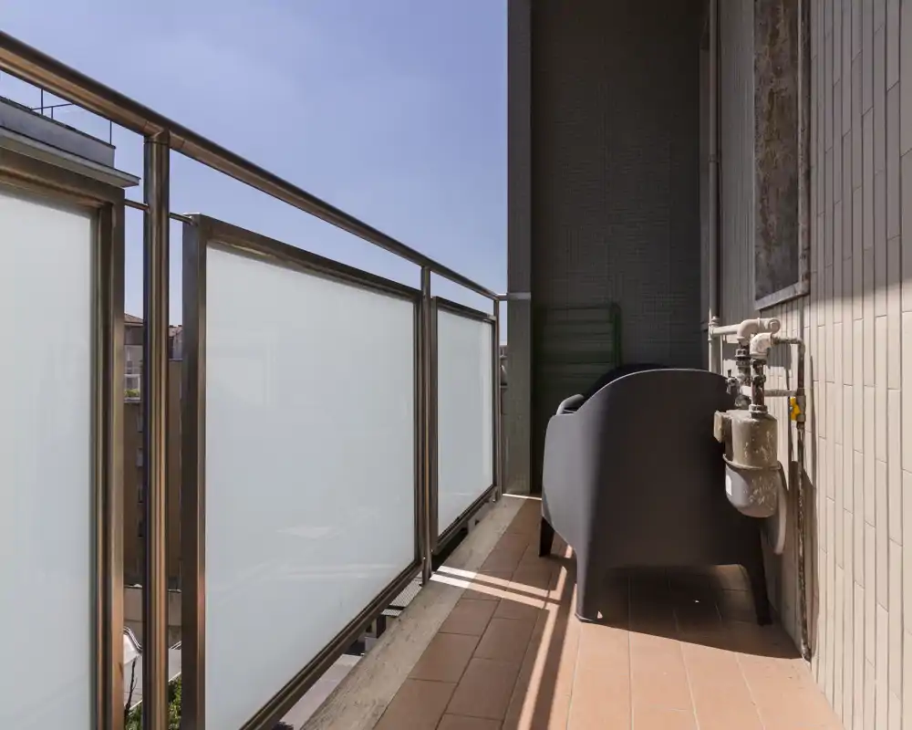 Balcone soleggiato e stretto con parapetto in vetro satinato, pavimento in piastrelle e una poltrona compatta — ideale per un caffè mattutino.