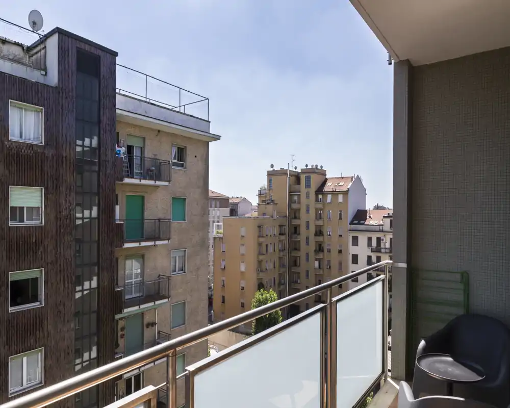 Balcone soleggiato con parapetto in vetro e vista sulla città; due sedute moderne e uno spazio compatto ideale per il caffè del mattino.