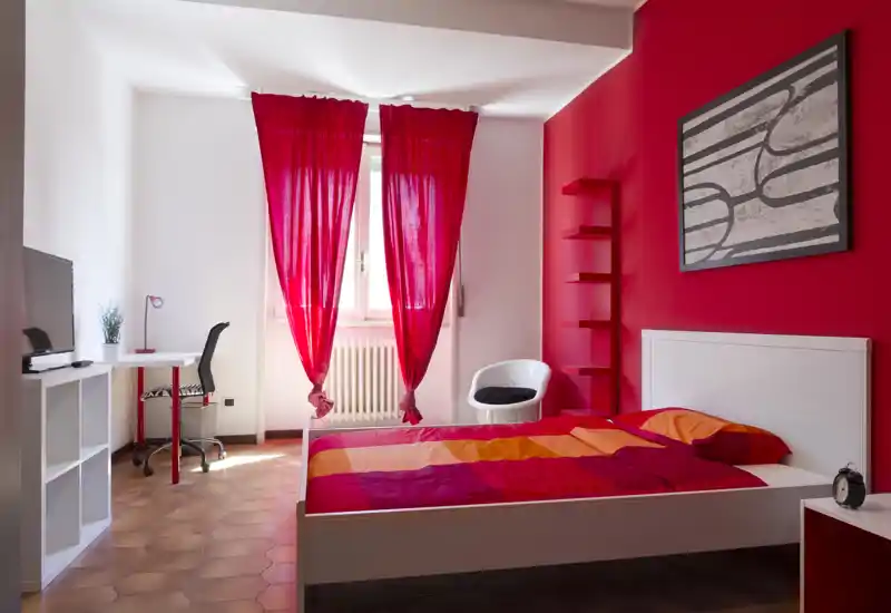 Camera da letto luminosa e moderna con struttura del letto bianca, biancheria dai toni rosso-arancio e parete accent rossa; la luce naturale dalla finestra e una scrivania piccola rendono lo spazio funzionale.