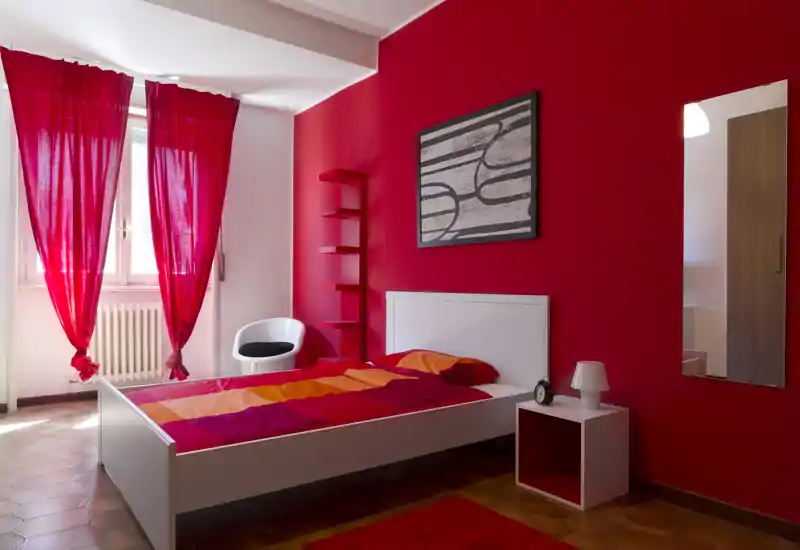 Camera da letto luminosa e di design con letto bianco, copriletto colorato e parete rossa a contrasto; la luce naturale dalla finestra rende l'ambiente arioso e accogliente.