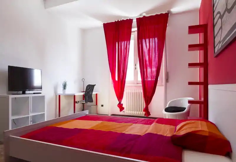 Camera da letto luminosa e colorata con un letto matrimoniale con biancheria rossa e arancione, finestra con tende rosse, una piccola scrivania e TV — moderna, ben illuminata e accogliente.