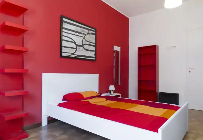 Camera da letto moderna e luminosa con struttura del letto bianca e biancheria rossa-arancione su una parete d'accento rossa; scaffalature minimal e uno specchio a tutta altezza completano l'arredamento semplice e di stile.