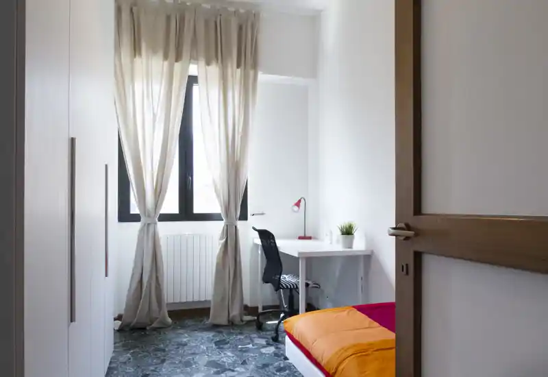 Bright, simply furnished bedroom with a single bed dressed in orange and red linens, a white desk with a chair and lamp by a large window with sheer curtains — ideal for a student or short-term rental.