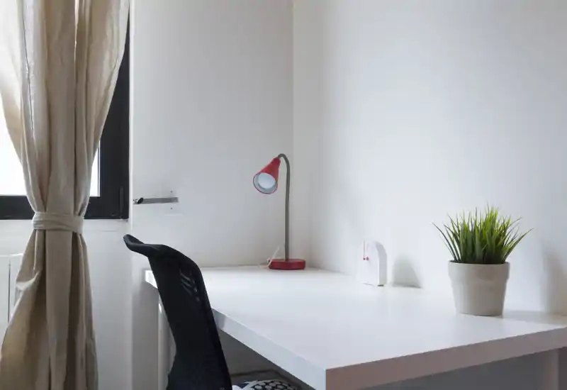 Bright, minimalist desk corner with a white worktable, ergonomic chair, red desk lamp and a potted plant—ideal for focused work or study.