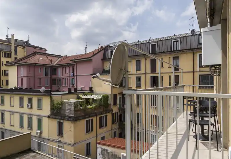 Sunny apartment balcony with metal railing, small table and chairs, and a view over neighboring buildings and rooftops.