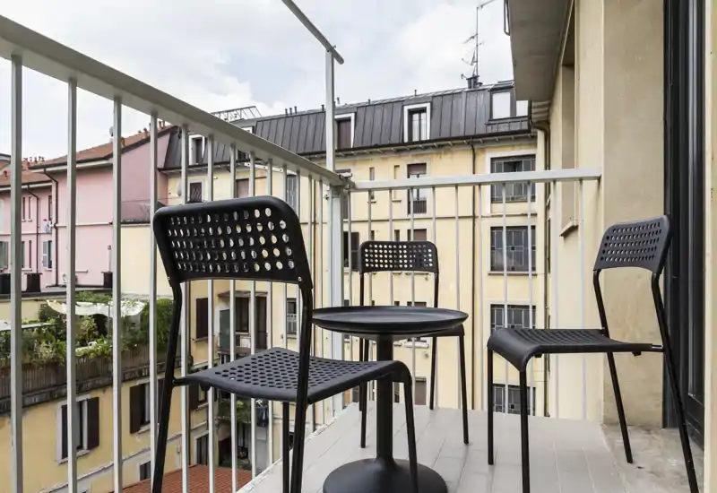 Balcony with a small round table and three perforated black chairs overlooking neighboring buildings, offering a compact outdoor seating area.