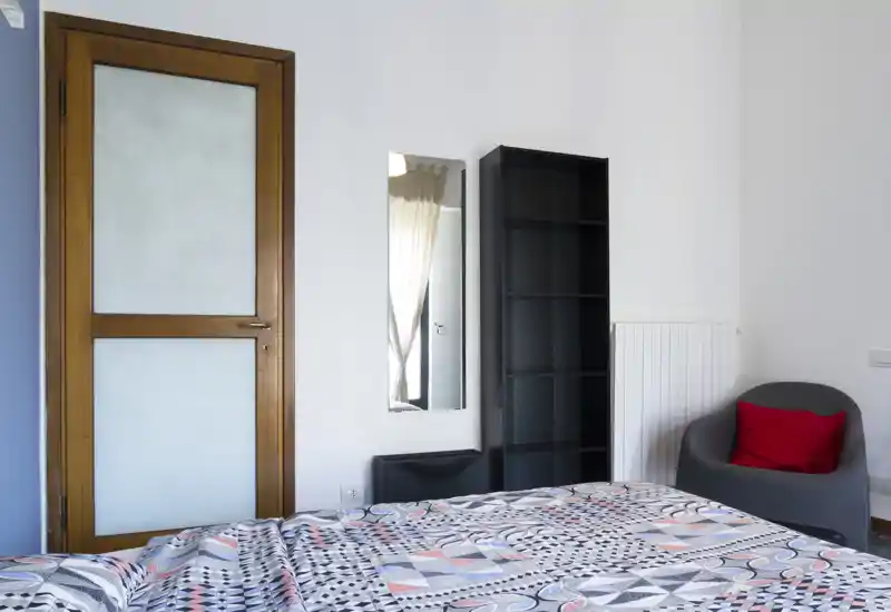 Bright, modern bedroom view showing a patterned bedspread in the foreground, a wooden door, wall mirror, shelving unit and an armchair with a red cushion.