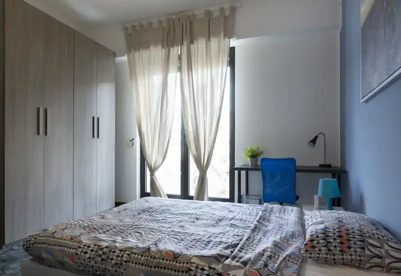 Bright, modern bedroom with a large patterned bed in the foreground, a tall wardrobe on the left and a desk with chair by the window letting in natural light.