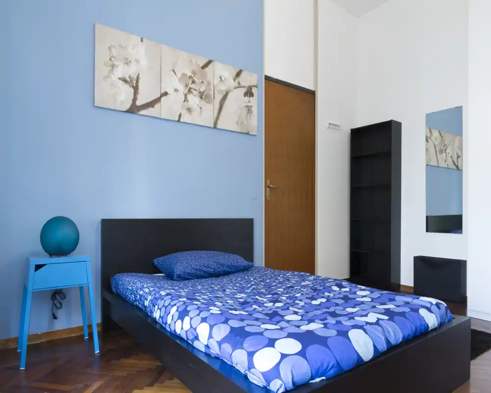 Bright, modern bedroom with a low black bed frame and blue patterned bedding. Pale blue accent wall, simple artwork above the headboard and a small bedside table with a spherical lamp create a calm, contemporary look.