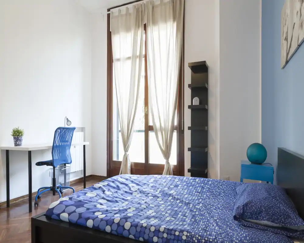 Bright, modern bedroom with a large bed dressed in a blue patterned duvet, natural light from tall windows, a small desk with chair and minimalist shelving.