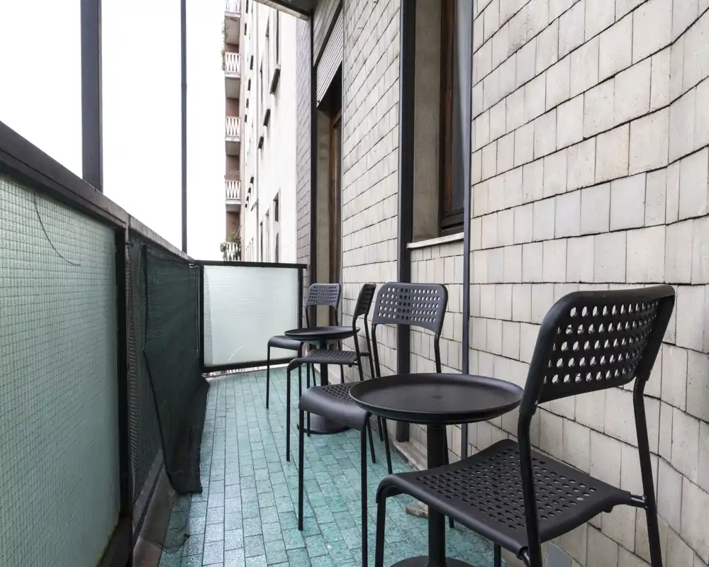 Sunny narrow balcony with turquoise tiled floor and a row of modern black perforated chairs and small round tables — ideal for coffee or light outdoor seating.