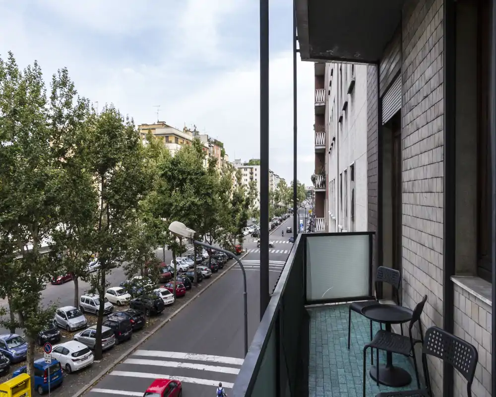 Street-facing balcony with two chairs and a small table; views over a tree-lined avenue and parked cars, suitable for morning coffee.