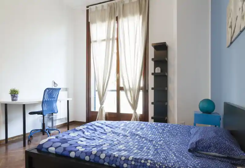 Camera da letto luminosa e moderna con un ampio letto con copriletto blu a motivi, molta luce naturale da grandi finestre, una scrivania con sedia e scaffalatura minimal.