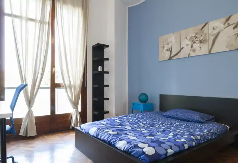 Camera da letto moderna e luminosa con letto matrimoniale, parete accentata azzurra e ampia finestra che lascia entrare molta luce naturale. Arredo semplice e biancheria a fantasia per un ambiente fresco e accogliente.