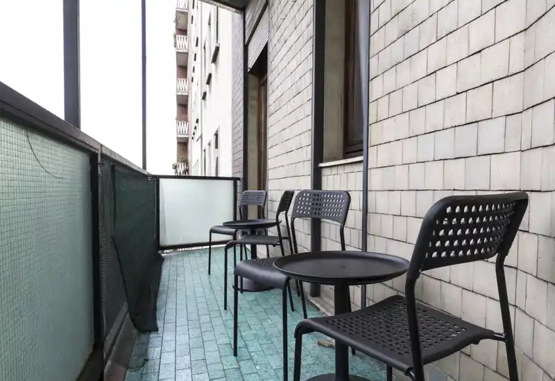 Balcone stretto con pavimento piastrellato turchese e una fila di moderne sedie nere traforate con piccoli tavolini rotondi — ideale per caffè o seduta all'aperto.