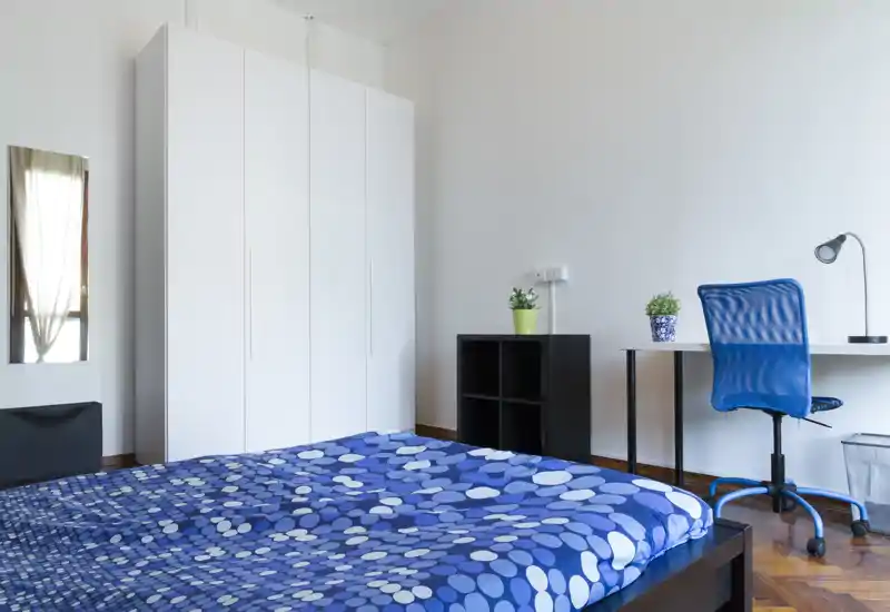Camera da letto luminosa e moderna con un grande letto blu a motivo, armadio bianco e una scrivania semplice con sedia blu. Linee pulite e arredo minimale la rendono fresca e funzionale per soggiorni brevi o lunghi.