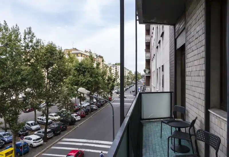 Balcone fronte strada con due sedie e un piccolo tavolo; vista su un viale alberato e auto parcheggiate, ideale per il caffè mattutino.