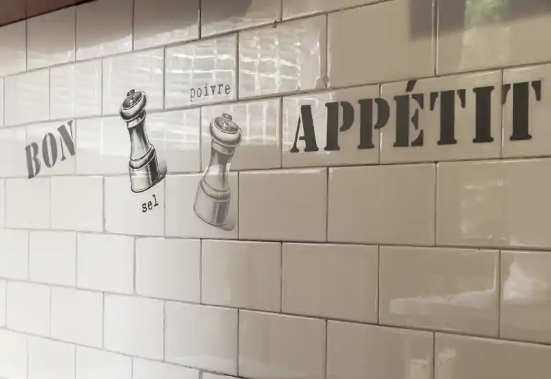 Primo piano di una parete della cucina piastrellata con la scritta decorativa 'Bon Appétit' e illustrazioni di macinini per sale e pepe; piastrelle lucide e riflessi naturali.