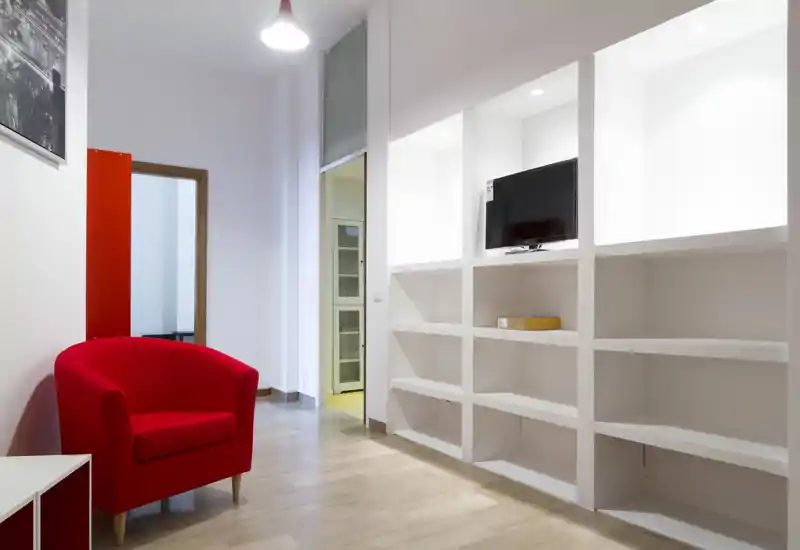 Piccolo soggiorno moderno con una poltrona rossa, scaffalature bianche incassate e TV a parete. Pulito, minimal e ben illuminato — adatto a un appartamento compatto.