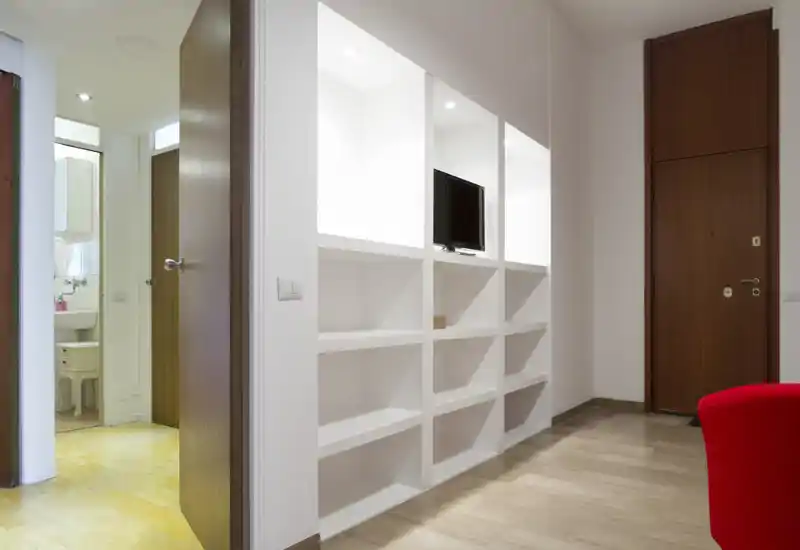 Ingresso luminoso e moderno con scaffalature bianche a scomparti integrate e porte in legno; pavimento in legno lucidato e una poltrona rossa in evidenza.