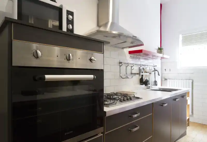 Cucina moderna e luminosa con forno in acciaio, piano cottura a gas, cappa e paraschizzi piastrellato bianco. Spazio compatto con cassetti, lavello sotto la finestra e una piccola mensola decorativa.