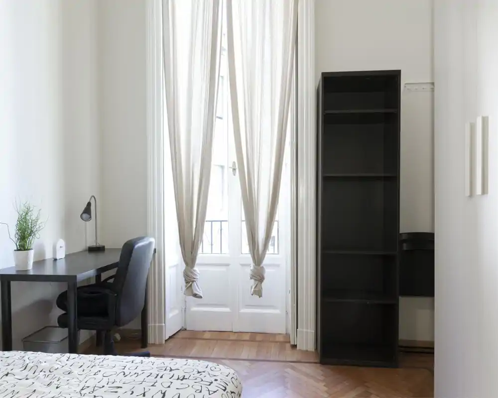 Coin de chambre lumineux et minimaliste avec lit, bureau et chaise près d'une porte-balcon ensoleillée, et une grande étagère ouverte — idéal pour une location aérée et rangée.