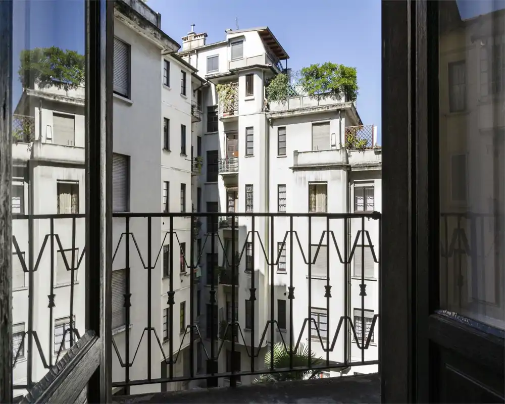 Petit balcon en ville encadré par une balustrade en fer décorative, donnant sur des immeubles voisins et un ciel bleu lumineux.