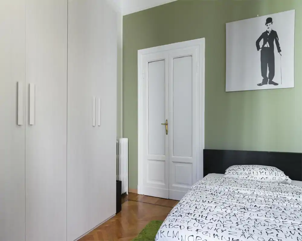 Chambre lumineuse et minimaliste avec une couette à motif, une tête de lit foncée et une grande armoire blanche. Le mur vert et le tableau apportent une ambiance moderne et apaisante.